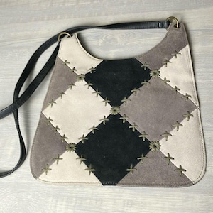 May include: A shoulder bag featuring a patchwork design of suede squares in black, light gray, and off-white. The squares are joined by visible cross-stitching in a light olive thread. The bag includes a thin black leather strap attached by rings.