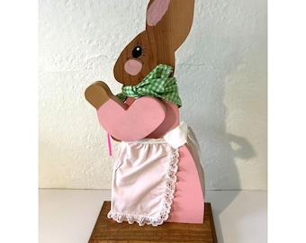 May include: A wooden rabbit figurine standing on a rectangular base. The rabbit is painted brown with pink inner ears and cheeks, wearing a green and white gingham neckerchief, a pink long-sleeved top, and a white apron with lace trim.