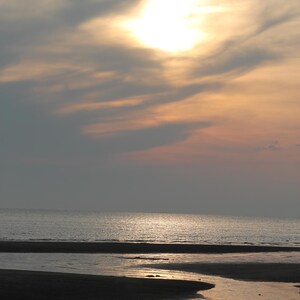 May include: A serene beach scene at sunset. The sky is filled with soft clouds, illuminated by the sun's glow. The ocean stretches to the horizon, reflecting the warm colors of the sky. The foreground shows wet sand and a small stream.
