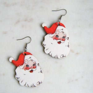 May include: A pair of Christmas-themed Santa Claus earrings. Each earring depicts a vintage-style Santa face with a red hat, white beard, and rosy cheeks. The earrings are made of wood and have silver-coloured fish hook ear wires.