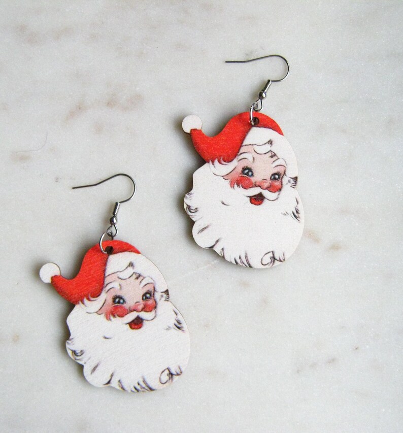 May include: A pair of Christmas-themed Santa Claus earrings. Each earring depicts a vintage-style Santa face with a red hat, white beard, and rosy cheeks. The earrings are made of wood and have silver-coloured fish hook ear wires.