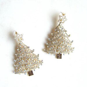 May include: A pair of gold tone earrings shaped like Christmas trees, each tree is covered in clear iridescent rhinestones.