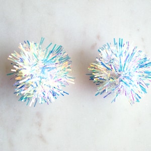 May include: Two iridescent white pom-poms with blue and silver accents. The pom-poms are made of thin, shiny strands of material.