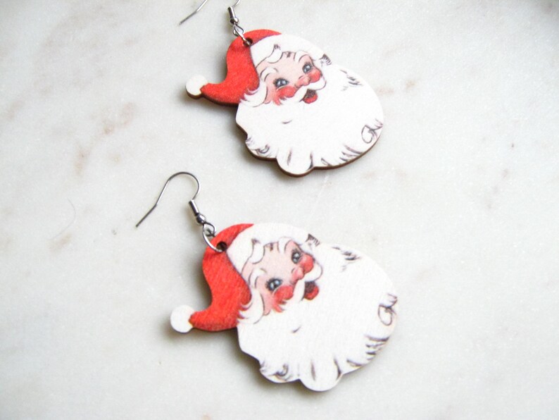 May include: A pair of festive earrings depicting Santa Claus. Each earring showcases Santa's face, complete with a red hat, white beard, and rosy complexion. The earrings are finished with silver-coloured ear wires.