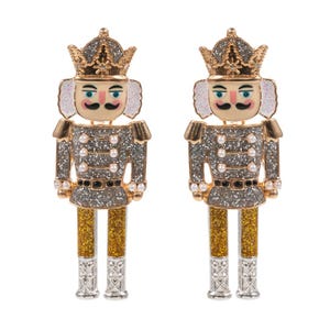 May include: A pair of festive nutcracker earrings. Each earring features a detailed design with a gold crown, a beige face with a black mustache, and a silver and gold uniform adorned with faux pearls. The legs are gold and silver.