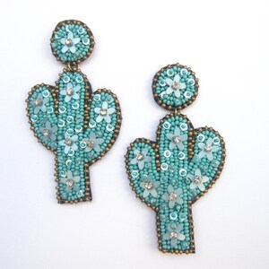 May include: A pair of turquoise blue cactus-shaped earrings with white flower accents. The earrings are made of beads and sequins.