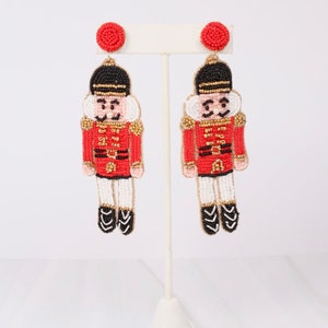 May include: A pair of beaded dangle earrings featuring a red and black nutcracker soldier design. The earrings have a red circular stud and a detailed depiction of the nutcracker in red, black, white, and gold. The earrings are displayed on a white earring stand.