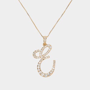 May include: A gold necklace with a pendant shaped like the letter "E". The pendant is set with numerous small, clear gemstones. The chain is a fine, gold-coloured box chain. The background is a plain, neutral colour.