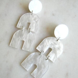 May include: A pair of white, abstract earrings. Each earring has a white circular stud, connected to two arch-shaped pieces. The earrings have a marbled, pearlescent effect, and are connected with silver-coloured jump rings. The earrings are displayed on a white marble surface.