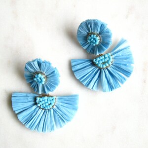 May include: Pale blue raffia earrings with a fan design and small turquoise beads. The earrings have a gold-coloured metal accent and a flower-like top. A unique accessory.
