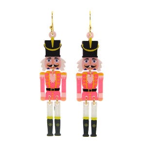 May include: A pair of dangling earrings featuring a pink and black nutcracker soldier design. The earrings have gold-colored hooks and pink and white accents. The nutcrackers have black hats, white mustaches, and pink uniforms.