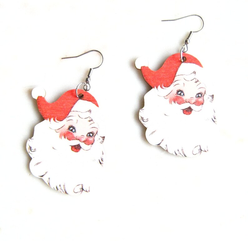 May include: A pair of Santa Claus earrings. Each earring depicts a smiling Santa face with a red hat, rosy cheeks, and a white beard. The earrings are made of a lightweight material and have silver-coloured fish hook ear wires.