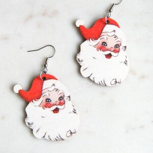 May include: A pair of Santa Claus earrings. Each earring features a red Santa hat, rosy cheeks, and a white beard. The earrings are made of wood and have silver-coloured fish hook ear wires. The earrings are designed for pierced ears.
