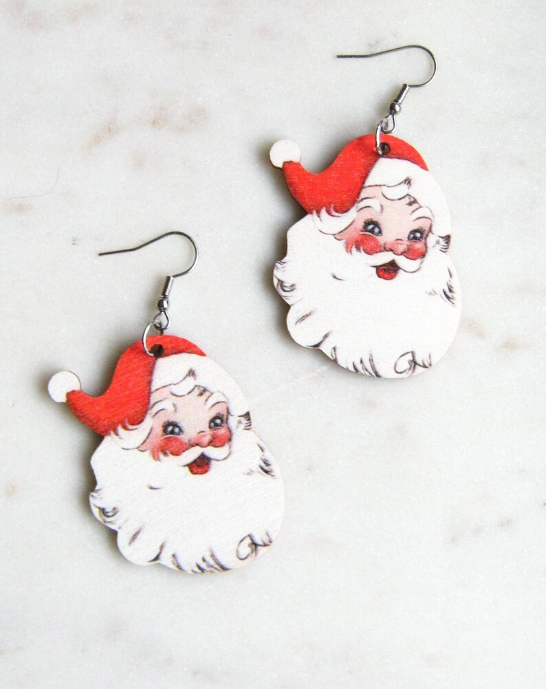 May include: A pair of Santa Claus earrings. Each earring features a red Santa hat, rosy cheeks, and a white beard. The earrings are made of wood and have silver-coloured fish hook ear wires. The earrings are designed for pierced ears.