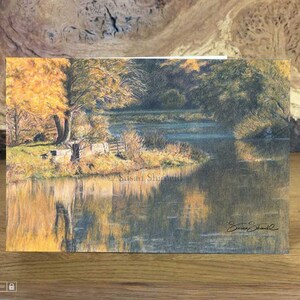 Puede incluir: Una pintura de acuarela de una escena de río con árboles a ambos lados del río. Los árboles están en tonos de verde y amarillo, y el agua es de un azul claro. La pintura está firmada "Susan Shield" en la esquina inferior derecha.