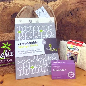 May include: A selection of sustainable cleaning products. The image shows compostable sponge cloths with a geometric design, a scrub pad, a washing-up pad, and a lavender soap bar. The items are arranged on a wooden surface.