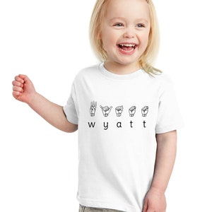 May include: White toddler t-shirt with the name Wyatt spelled out in British Sign Language. The letters are represented by hand gestures.