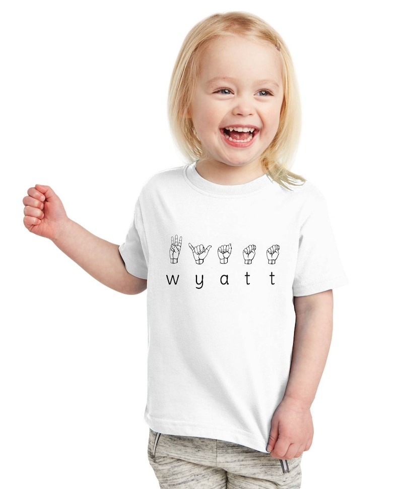 May include: White toddler t-shirt with the name Wyatt spelled out in British Sign Language. The letters are represented by hand gestures.