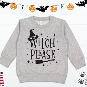 Può includere: Una felpa grigia con un cappello da strega nero, una scopa e il testo "Witch Please" stampato sul davanti. La felpa è decorata con piccole stelle nere.