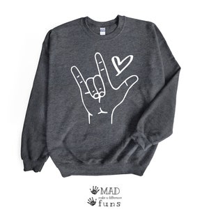 May include: A dark grey sweatshirt with a white graphic of a hand signing "I love you" in American Sign Language. A small heart is above the hand.