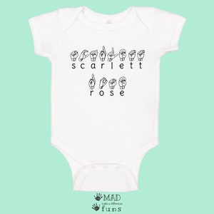 May include: White baby onesie with the name "Scarlett Rose" spelled out in British Sign Language. The onesie has short sleeves and a snap closure.