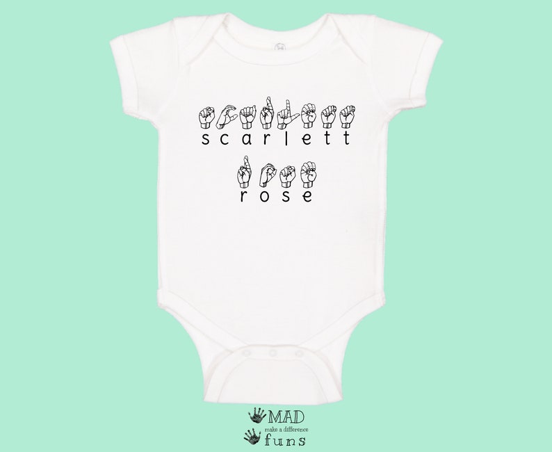 May include: White baby onesie with the name "Scarlett Rose" spelled out in British Sign Language. The onesie has short sleeves and a snap closure.