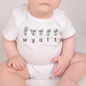 May include: White baby onesie with the name "Wyatt" spelled out in black letters and the letters signed in British Sign Language.