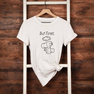 May include: White t-shirt featuring the text "But First," above a black graphic of two fists clenching, with a recycling symbol between them. The shirt is displayed on a wooden hanger against a wooden backdrop.