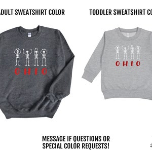 May include: Two sweatshirts, one adult and one toddler size, with a design of four skeletons with their arms raised in the "O-H" pose and the word "OHIO" underneath. The adult sweatshirt is dark grey and the toddler sweatshirt is light grey. Both sweatshirts have red lettering.