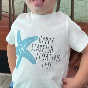 May include: A white t-shirt with a blue starfish graphic and the text "HAPPY STARFISH FLOATING FREE". A young child is wearing the shirt and sticking their tongue out.
