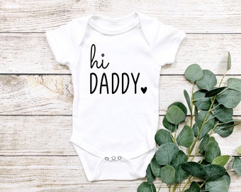 May include: White baby onesie with black text that reads "hi daddy" with a small black heart.