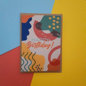 May include: A colourful birthday card featuring abstract shapes in shades of blue, green, yellow, orange and red. The card reads "Happy Birthday!" in a white font.