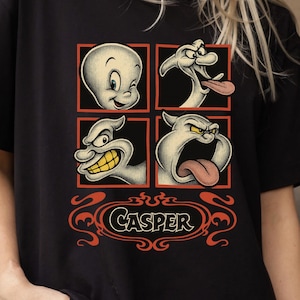May include: Black t-shirt with a four-panel graphic of Casper the Friendly Ghost displaying various expressions. The Casper logo is in an orange decorative design at the bottom. The shirt is made of a soft material.