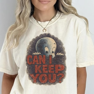 May include: Cream-coloured t-shirt with a vintage graphic of Casper the Friendly Ghost. The design includes the text "CAN I KEEP YOU?" in red letters, set against a brown, textured background. The shirt has short sleeves.