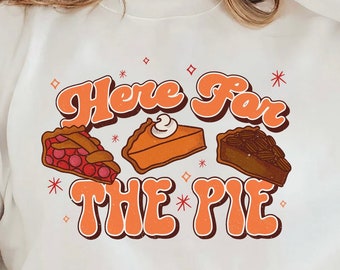 Puede incluir: Sudadera blanca con el texto "Here For The Pie" en naranja. El gráfico incluye ilustraciones de tres porciones de tarta: cereza, calabaza y nuez. El diseño tiene un estilo retro.