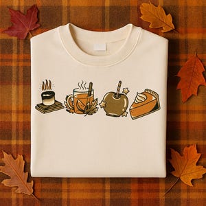 Op de afbeelding: Crèmekleurige sweatshirt met een herfstthema. Het ontwerp omvat een s'more, een mok cider met een kaneelstokje, een karamelappel en een stuk pompoentaart. De sweatshirt is omgeven door herfstbladeren.