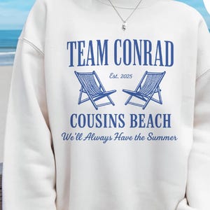 Op de afbeelding: Witte sweatshirt met blauwe tekst met de tekst "TEAM CONRAD" boven twee blauwe strandstoelen en "COUSINS BEACH" met de zin "We'll Always Have the Summer". De tekst "Est. 2025" is ook inbegrepen.