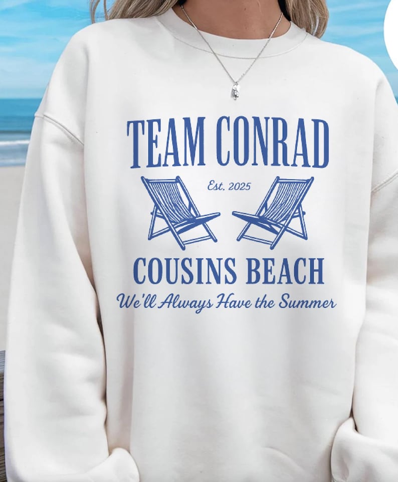 Op de afbeelding: Witte sweatshirt met blauwe tekst met de tekst "TEAM CONRAD" boven twee blauwe strandstoelen en "COUSINS BEACH" met de zin "We'll Always Have the Summer". De tekst "Est. 2025" is ook inbegrepen.