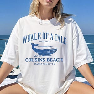 Op de afbeelding: Wit T-shirt met een blauwe walvisafbeelding en de tekst "WHALE OF A TALE BOOKSHOP COUSINS BEACH MASSACHUSETTS". Het shirt heeft een relaxte pasvorm en korte mouwen. Het ontwerp toont een walvisillustratie.