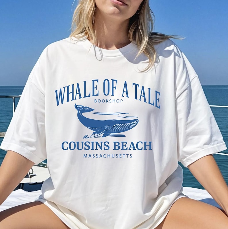 Op de afbeelding: Wit T-shirt met een blauwe walvisafbeelding en de tekst "WHALE OF A TALE BOOKSHOP COUSINS BEACH MASSACHUSETTS". Het shirt heeft een relaxte pasvorm en korte mouwen. Het ontwerp toont een walvisillustratie.