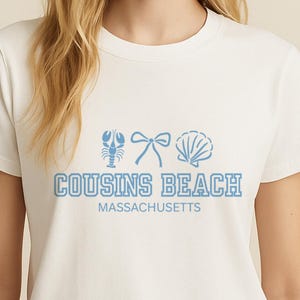 Op de afbeelding: Wit T-shirt met een lichtblauwe afbeelding van een kreeft, strik en schelp. De tekst "COUSINS BEACH MASSACHUSETTS" staat eronder in lichtblauw gedrukt. Ronde hals.