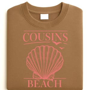 Op de afbeelding: Een gevouwen bruine sweatshirt met de roze tekst "COUSINS BEACH", een schelpgrafiek en vliegende vogels. De sweatshirt heeft een ronde hals en een blanco wit label.