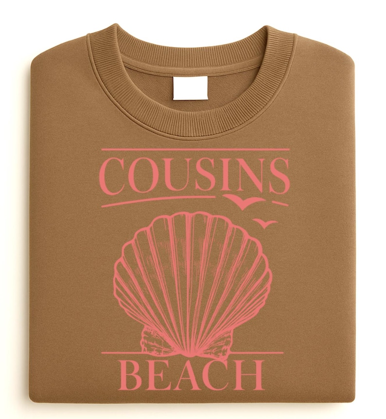 Op de afbeelding: Een gevouwen bruine sweatshirt met de roze tekst "COUSINS BEACH", een schelpgrafiek en vliegende vogels. De sweatshirt heeft een ronde hals en een blanco wit label.