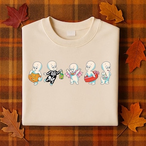 Puede incluir: Sudadera color crema con un dise&ntilde;o de Halloween. El dise&ntilde;o presenta cinco fantasmas de dibujos animados disfrazados, incluyendo un esqueleto, un fantasma con una calabaza y otros con capa, flotador y cesta. El dise&ntilde;o est&aacute; centrado en la parte delantera.