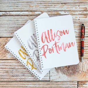 May include: Three white spiral notebooks with gold, silver, and red lettering. The top notebook has the name "Allison Portman" written in red.