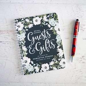 May include: A black and white floral patterned notebook with a spiral binding. The cover has the text "Guests & Gifts" in white cursive lettering. The text "Bridal Shower" is in smaller white lettering above the cursive text. The text "In Honor of Samantha Parker October 22, 2016" is in white lettering below the cursive text.