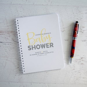 May include: A white spiral bound notebook with a gray and yellow baby shower announcement. The announcement says "Audrey & Harper's Baby Shower" and "Guests & Gifts in honor of Baby Charlotte, October 22, 2016".