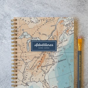 May include: A spiral bound notebook with a vintage map design featuring the eastern coast of the United States. The cover has the words "Adventures" and "rachel norlin" printed on it. A pencil is lying next to the notebook.