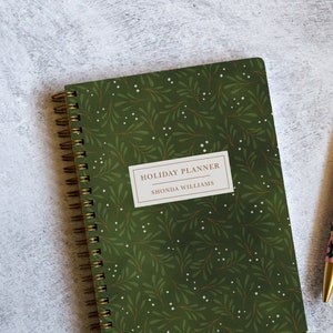 May include: A green spiral bound planner with a white and brown floral pattern and the text "HOLIDAY PLANNER SHONDA WILLIAMS" on the front cover. A gold pen with a floral pattern is lying on a gray surface next to the planner.