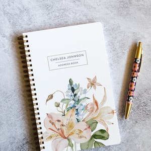 May include: A white spiral bound address book with gold rings. The cover features a watercolor floral design with pink, yellow, and blue flowers. The text "Chelsea Johnson Address Book" is printed in black on a white rectangle.